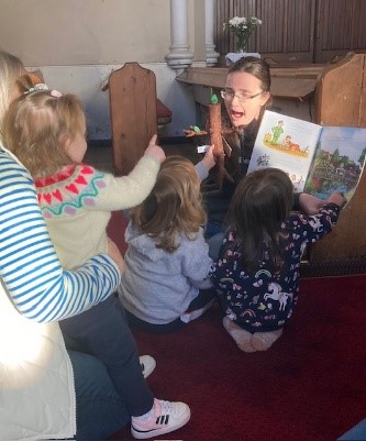 Story being read to a group of children. Some of them point to the book, another holds up a stickman toy