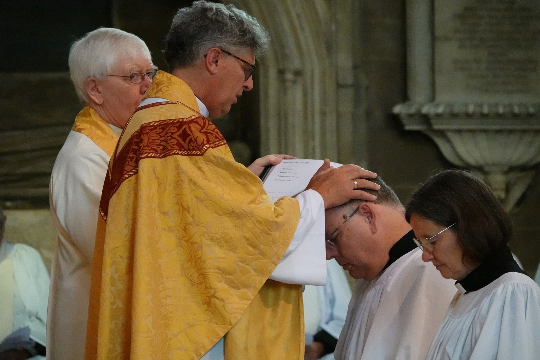Bishop Martin with his hands on Paul Latham's head as he ordains him. Paul kneels next to Philippa Lowe