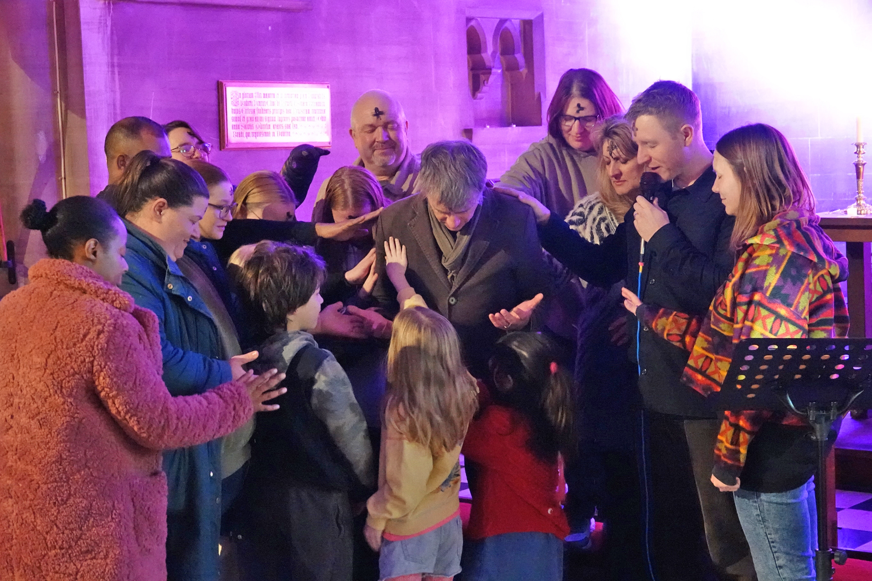 A group of people, both adults and children standing around Bishop Hugh as they pray for him.