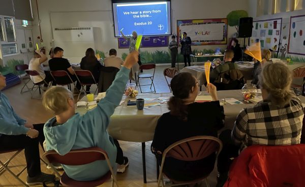 Members of the congregation are seated at tables facing the vicar. Some of them wave glow sticks during the bible story taken from Exodus 20.