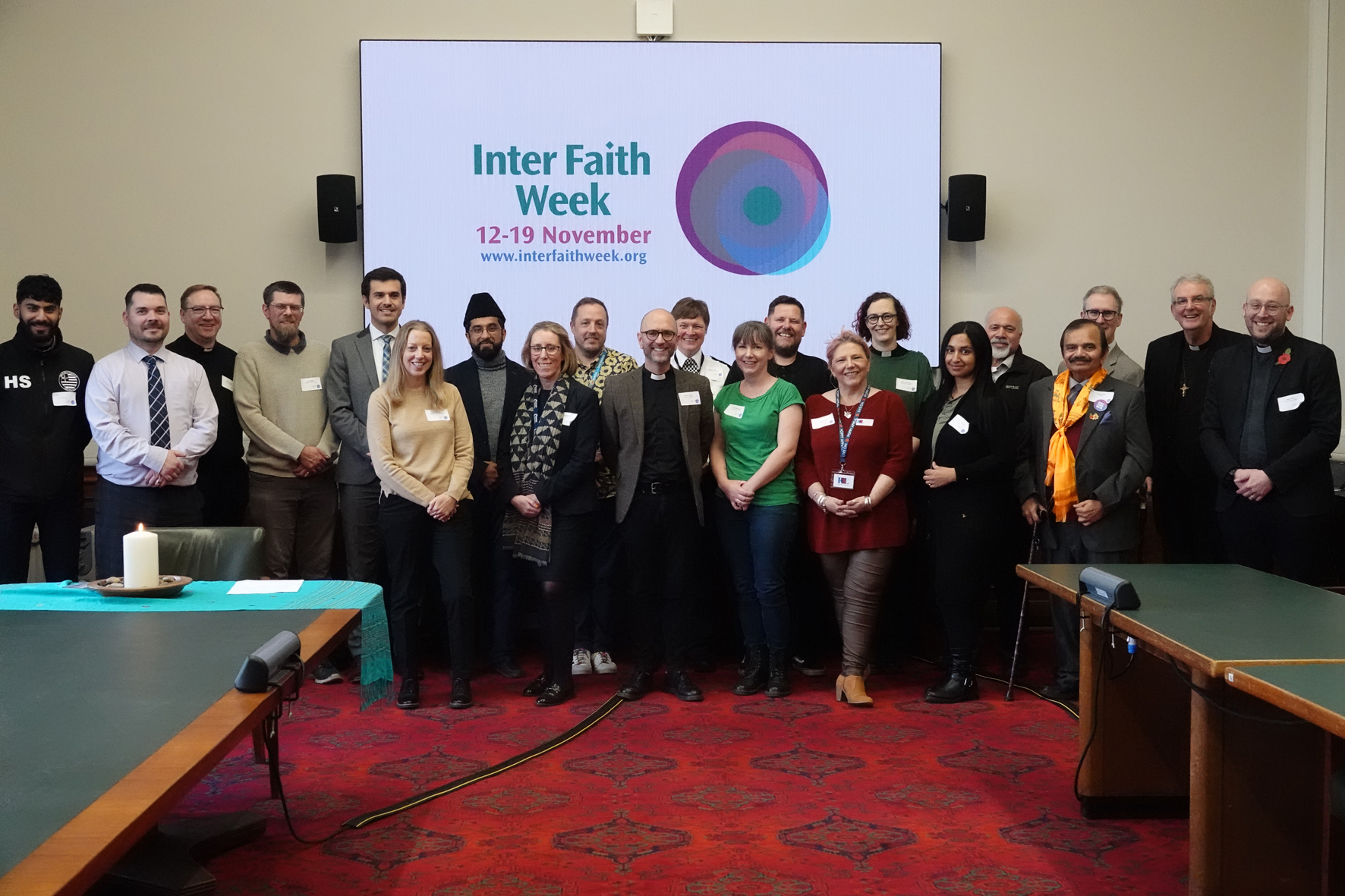 People gathered for the interfaith reception standing at the front of the room in front of an interfaith week sign