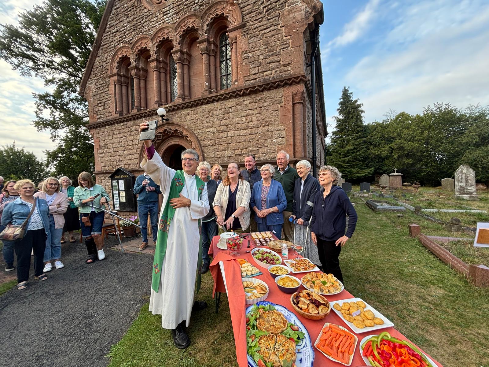 Bishop Martin takes a selfie with a group of people in front of Trimpley church while more people look on and a table laden with food is in the foreground