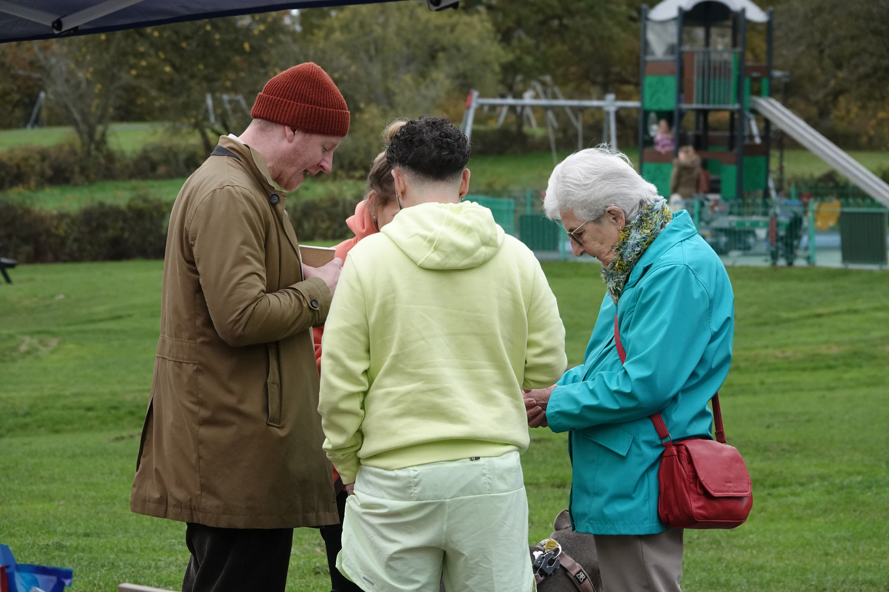Four people standing together outside in a park with their heads bowed in prayer