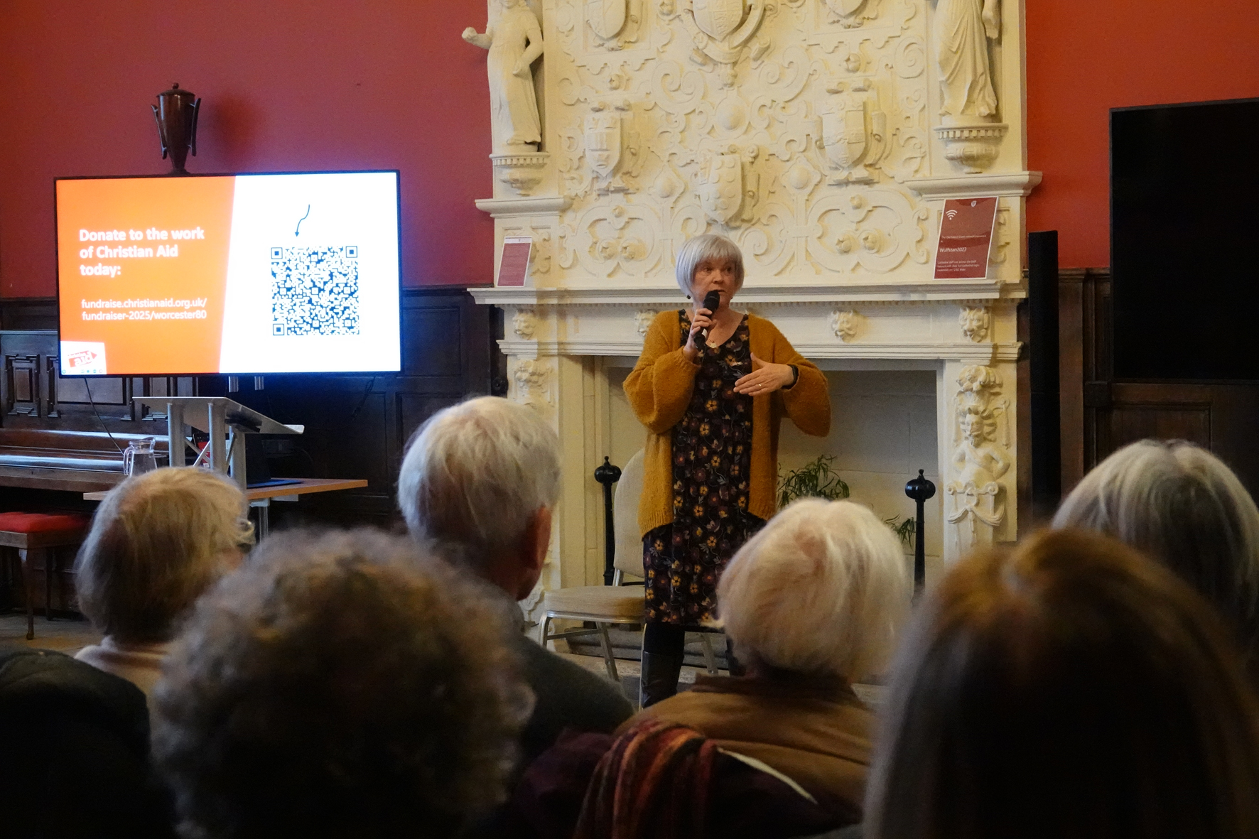 Alison standing in front of an audience with a microphone and next to a Christian Aid poster