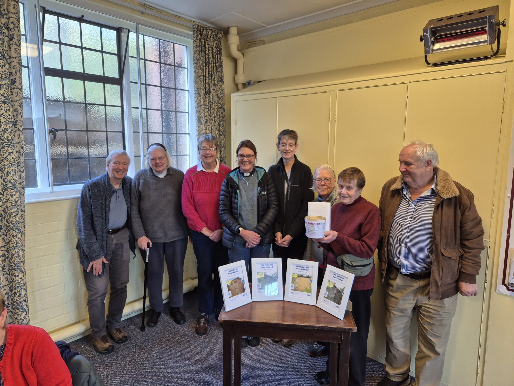 7 people standing in a semi circle in a room with the toilet twinning certificates in front of them on a table. 
