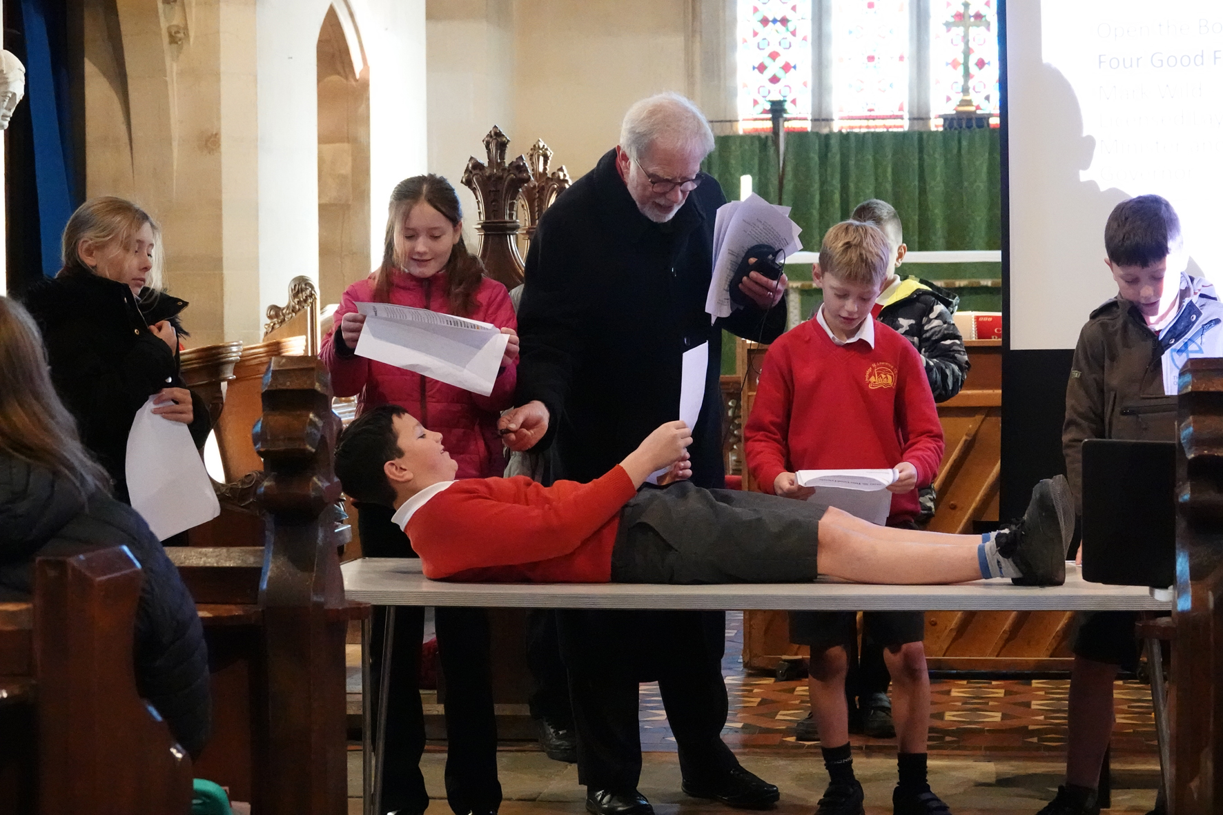 Children stood at the front of church holding scripts with one person laying on a table. An adult stands in the middle holding a microphone