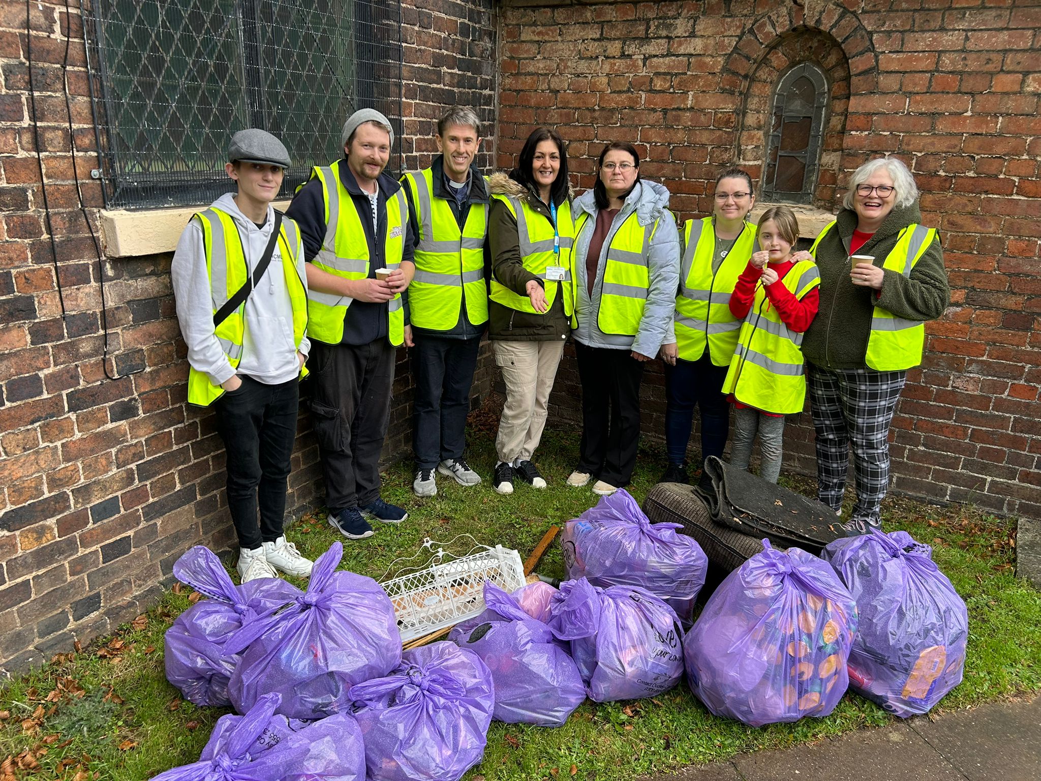 A group of people in hi-vis tabards sanding in front of the church with around 10 purple bin bags of rubbish in front of them.