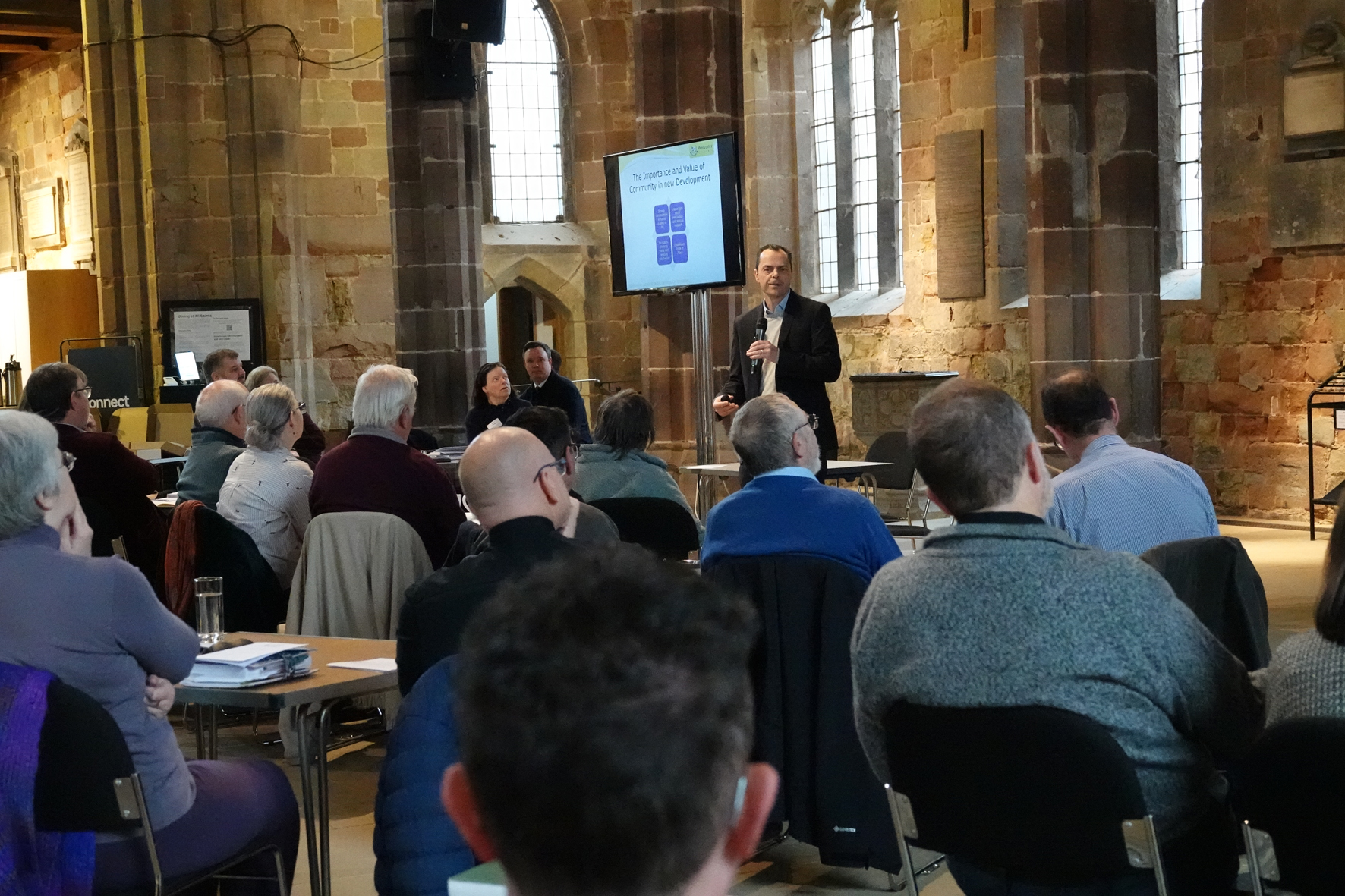 Photo taken from behind people sitting at tables and listening to Duncan Rudge who stands in front of a screen in St Helen's Church