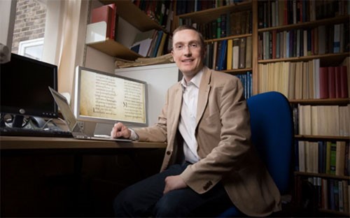 Hugh Houghton sitting at his desk looking at the camera with shelves of books behind and a manuscript on his screen