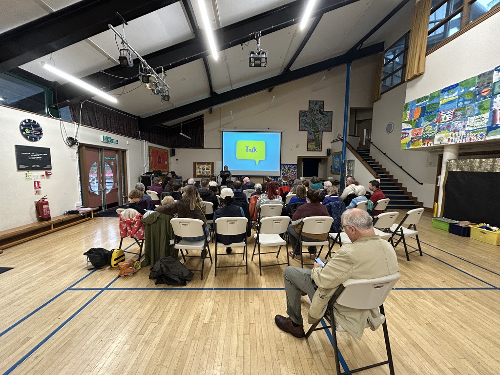 Photo taken from the back of a school hall where people sit in rows in front of a screen with 'talk' on it