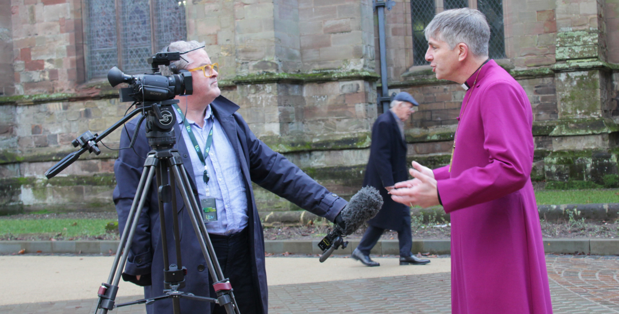 Bishop Hugh being interviewed by a journalist holding a microphone and standing next to a camera
