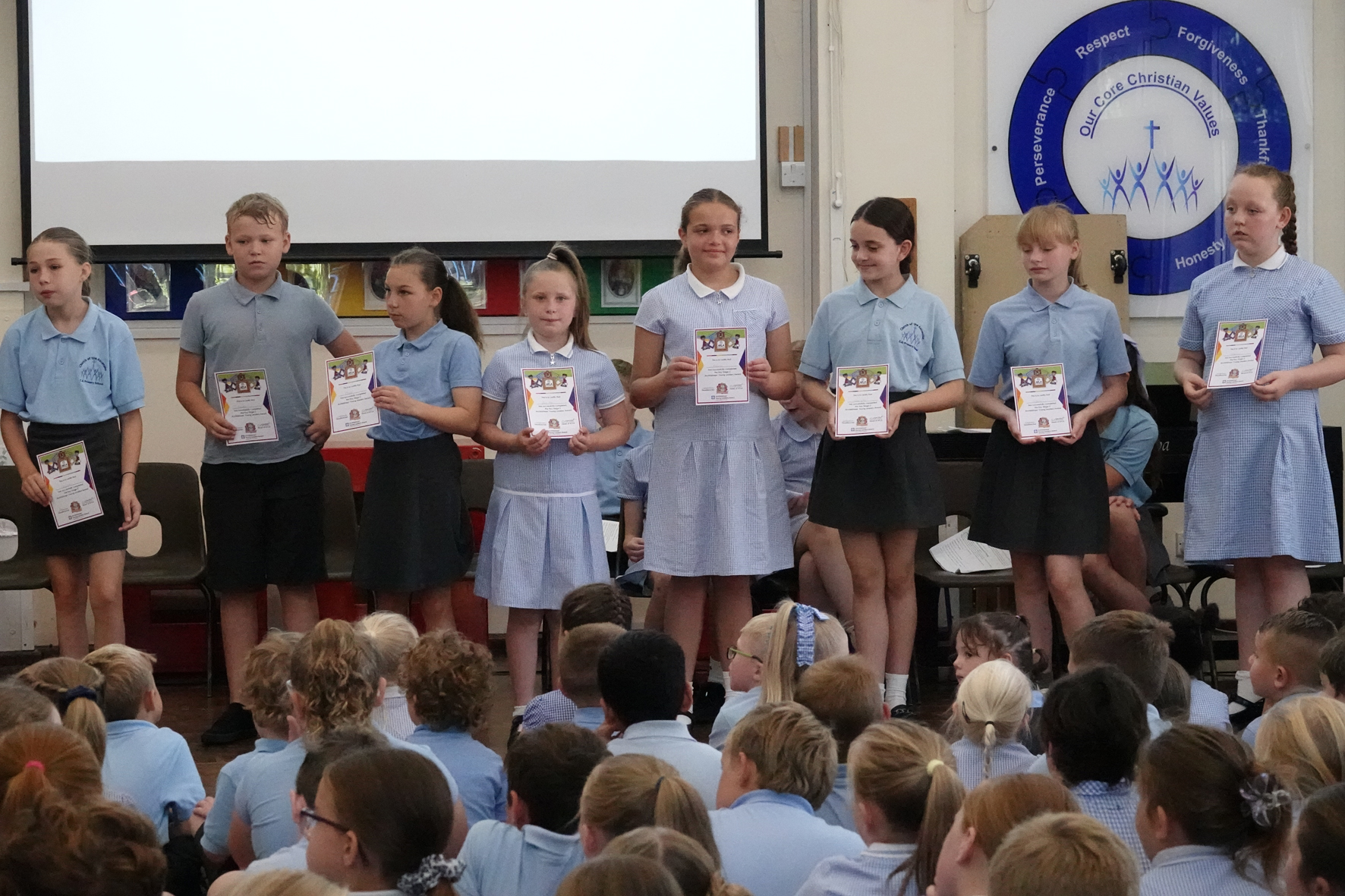 Children holding their certificates and standing at the front of assembly with children sitting in front of them