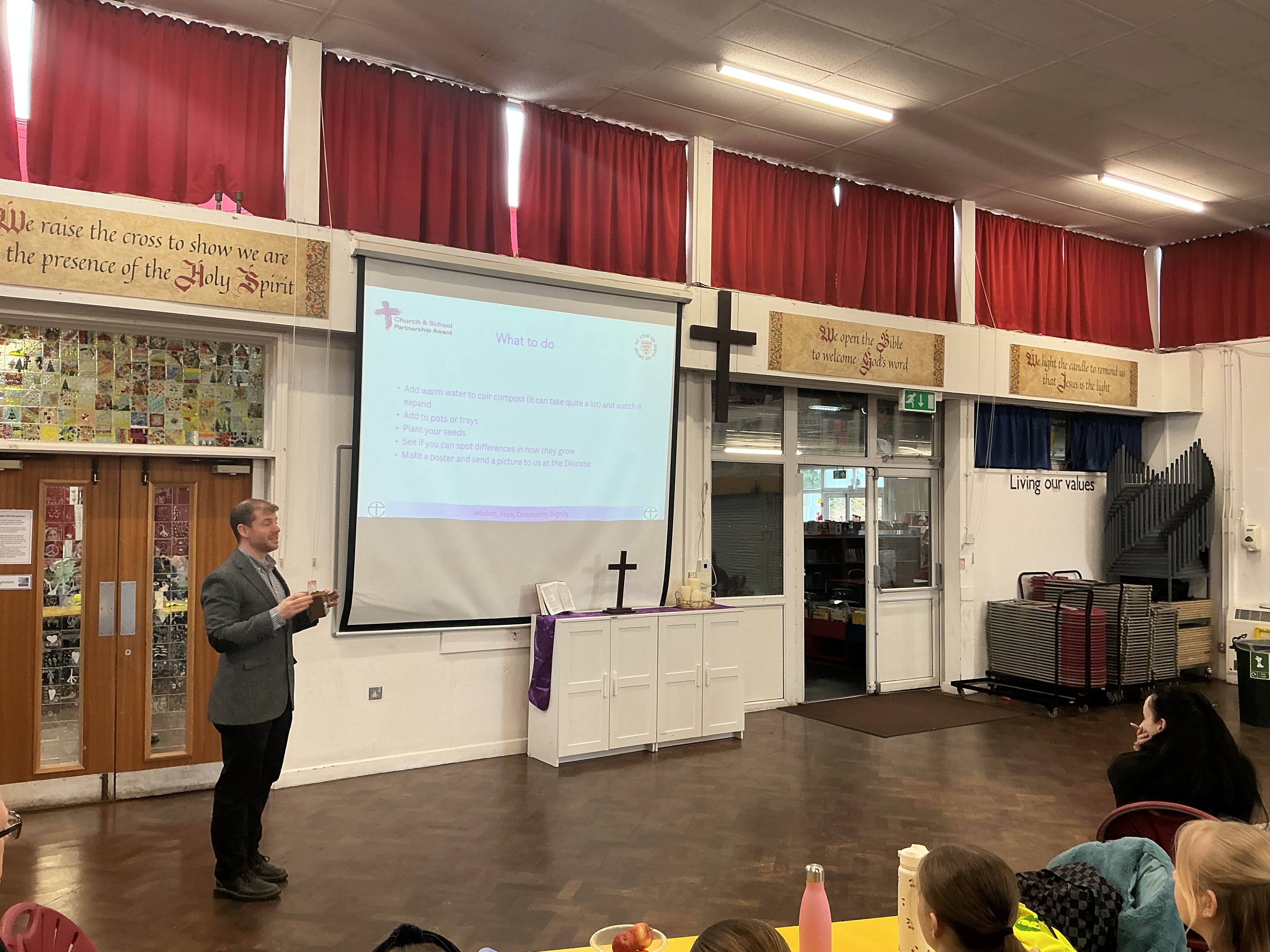 Lee standing in front of a big screen talking to children sitting in a school hall