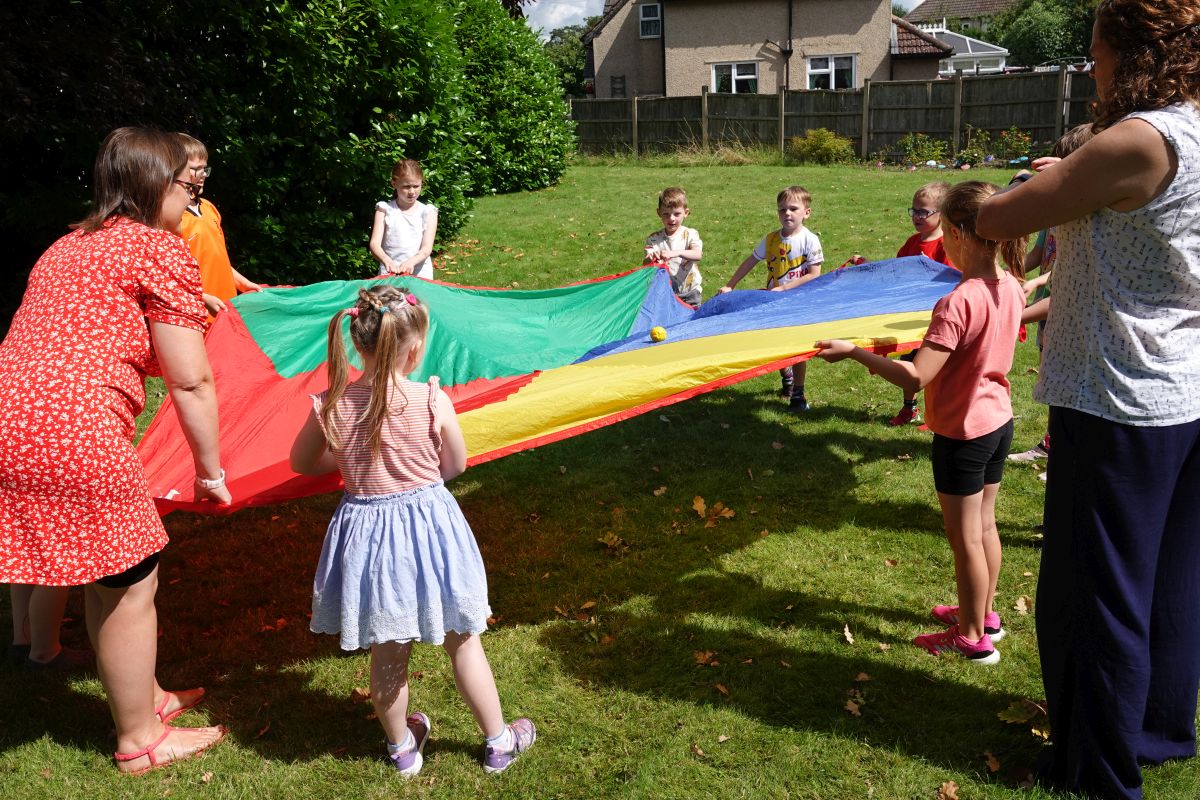 Supported by adults the children stand in a circle holding onto a brightly coloured parachute spread between them, trying to bounce a ball on top of it.