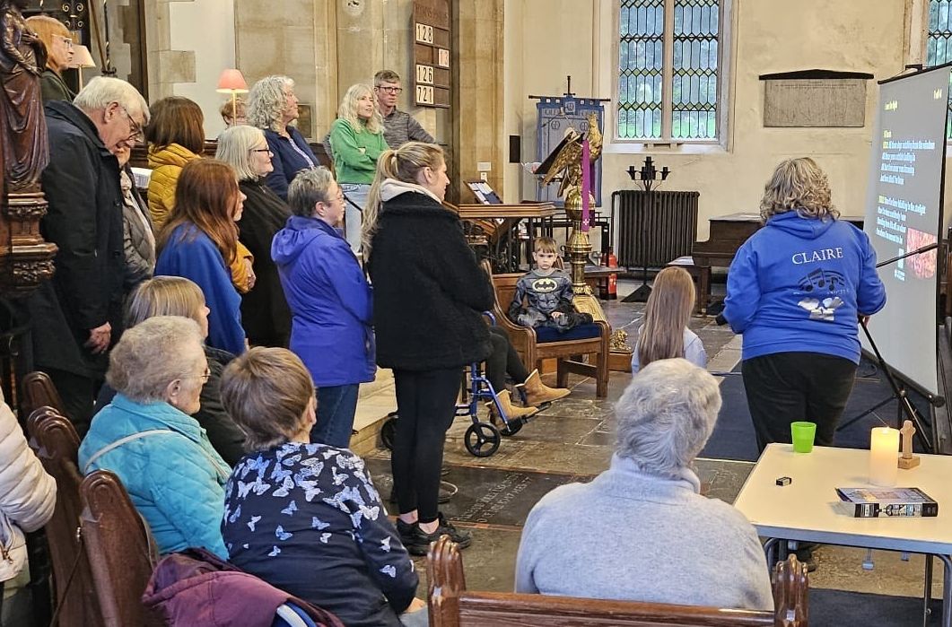 People of different ages gather round Claire and a screen with words on to sing