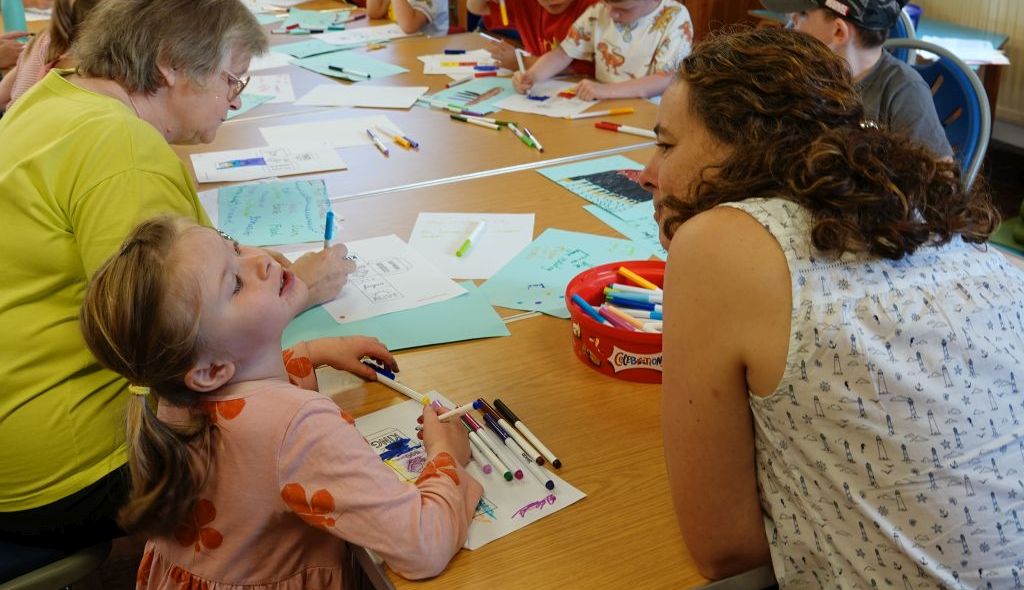 Helen Laird sits at a table with children and adults who are drawing and colouring