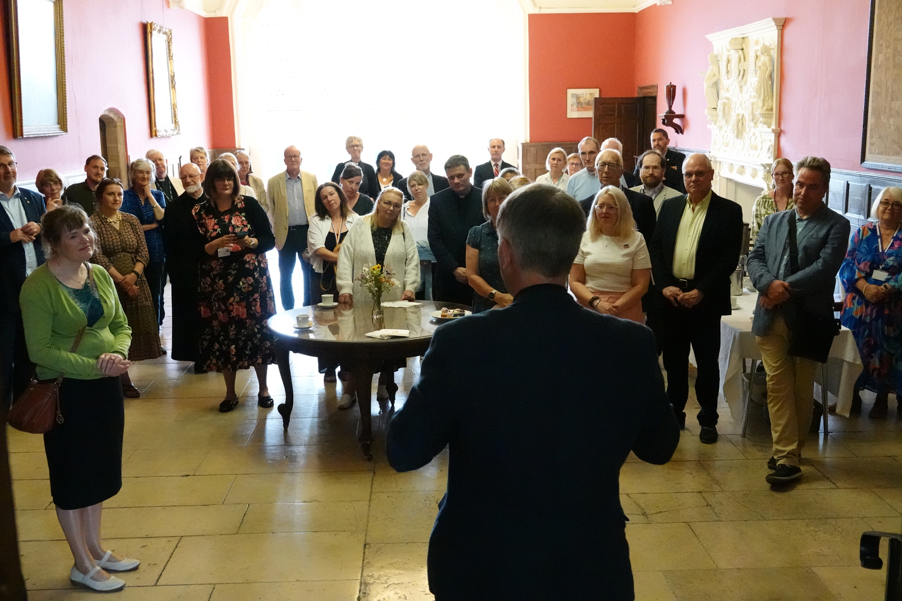 Looking towards a big window in the great hall, Bishop Hugh has his back to the camera as he talks to a group of gathered people