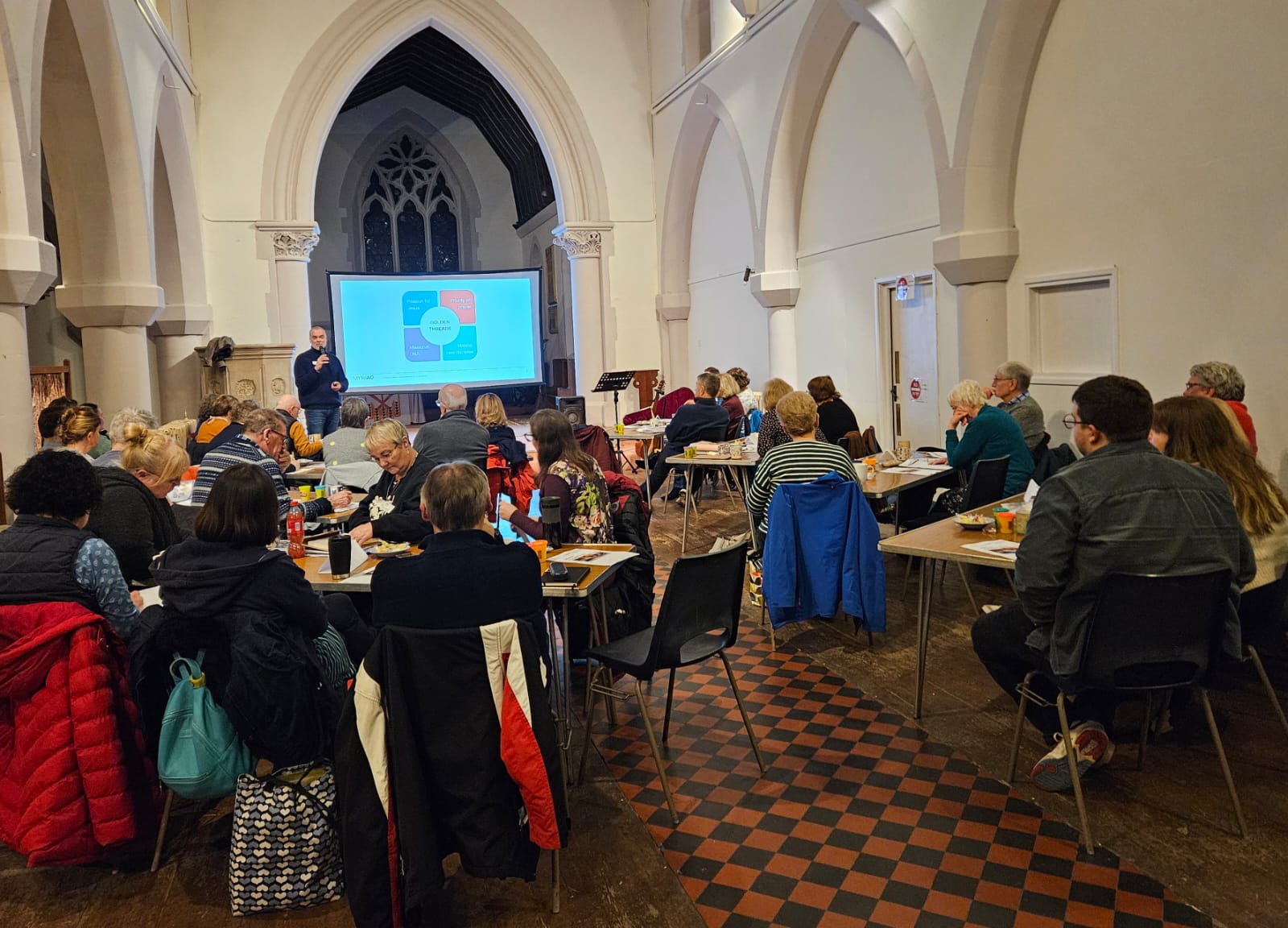 Photo from the back of the room with people in the NWC learning community sat around tables & looking at the screen at the front