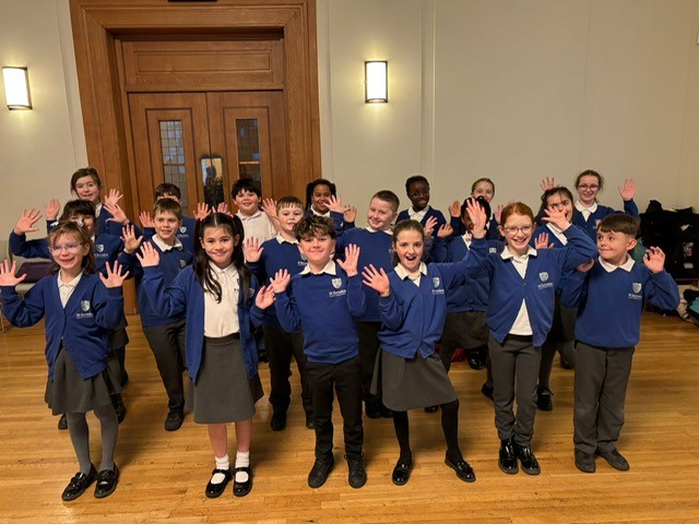 A group of children in St Barnabas school uniform standing in a hall and waving at the camera