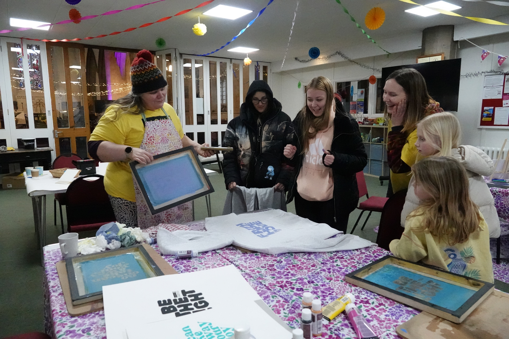 A group of people gathered around a table with a hoodie on the table onto which a phrase has just been screenprinted.