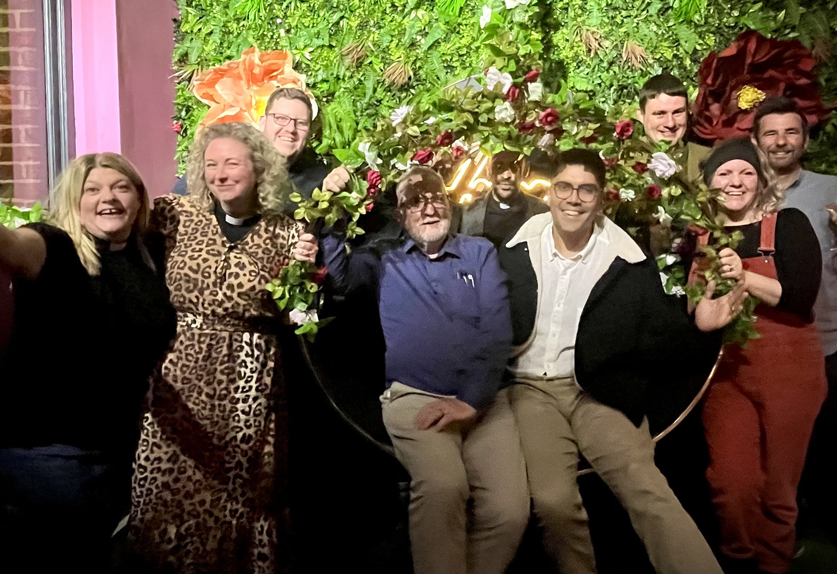 Joseph sitting with a group of smiling curates in front of a green botanical wall