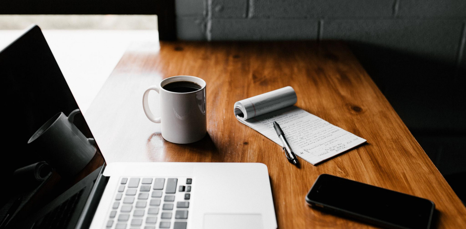 A laptop with a notebook and pen and cup of coffee. 